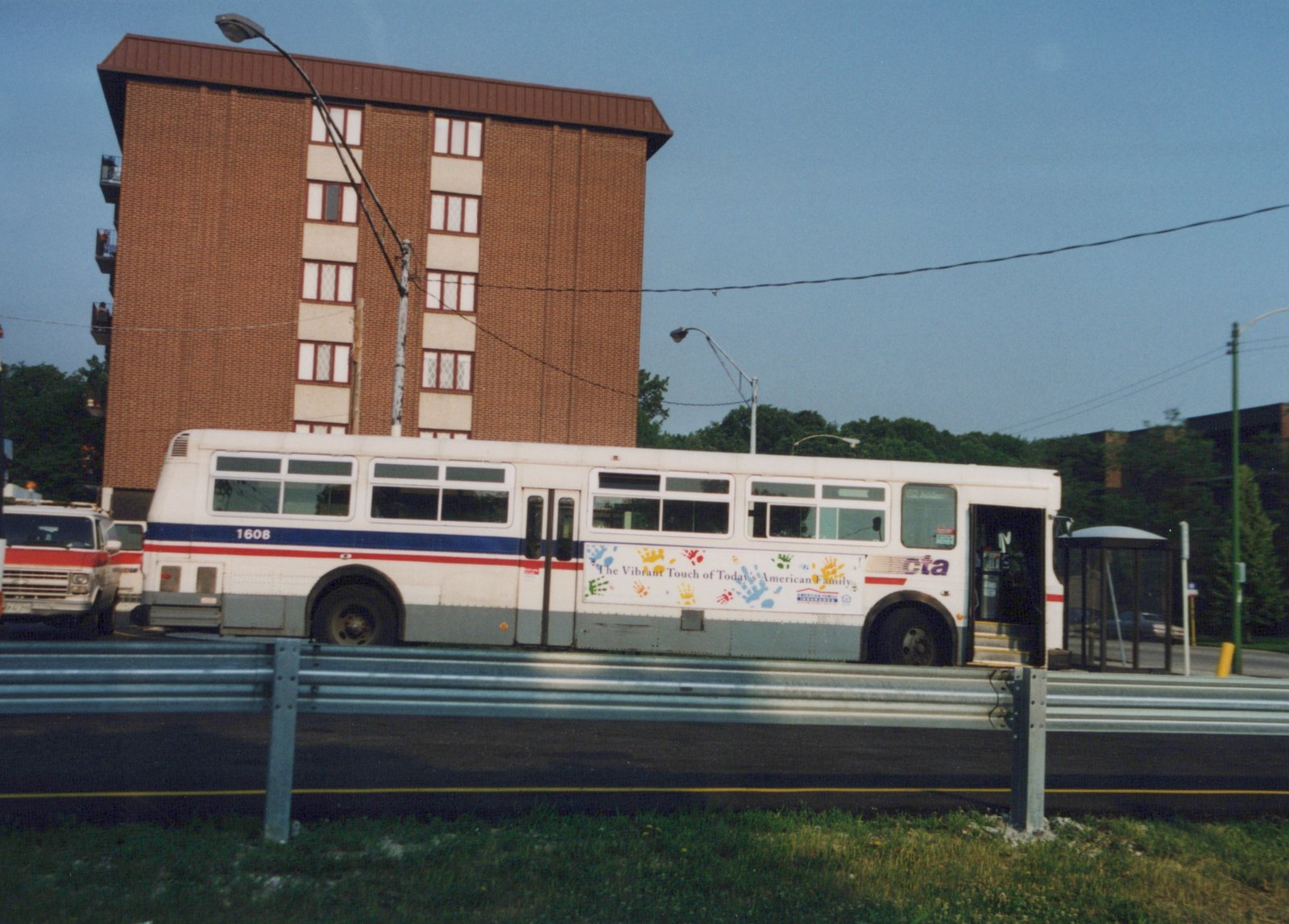 Side view of a white CTA bus with the number 1608, parked next to a guardrail with a red and white pickup truck behind it and a tall brick building in the background.