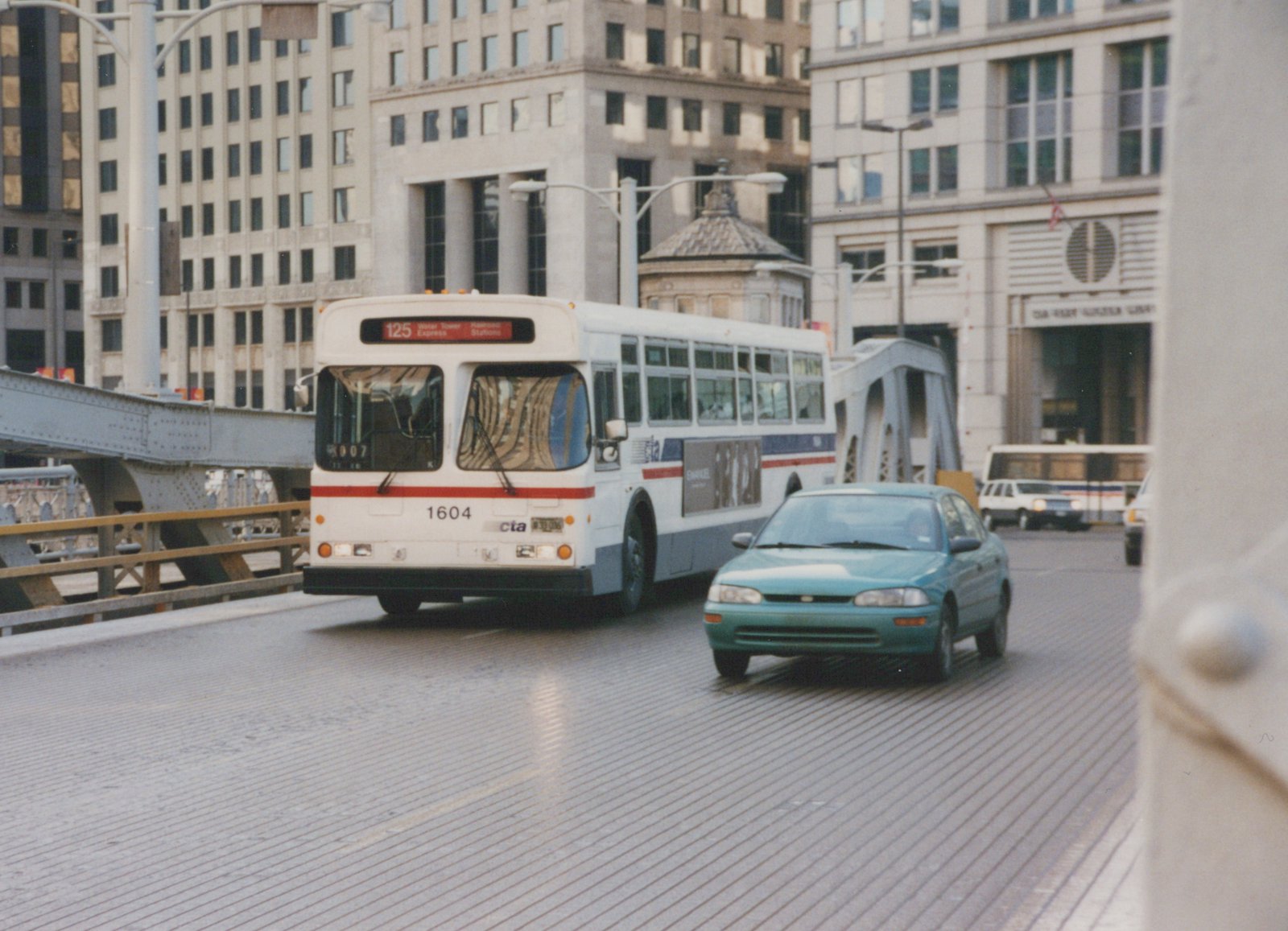 A white and blue Chicago Transit Authority bus, number 1604, is driving on a metal grate bridge with a teal car in front of it and city buildings in the background.