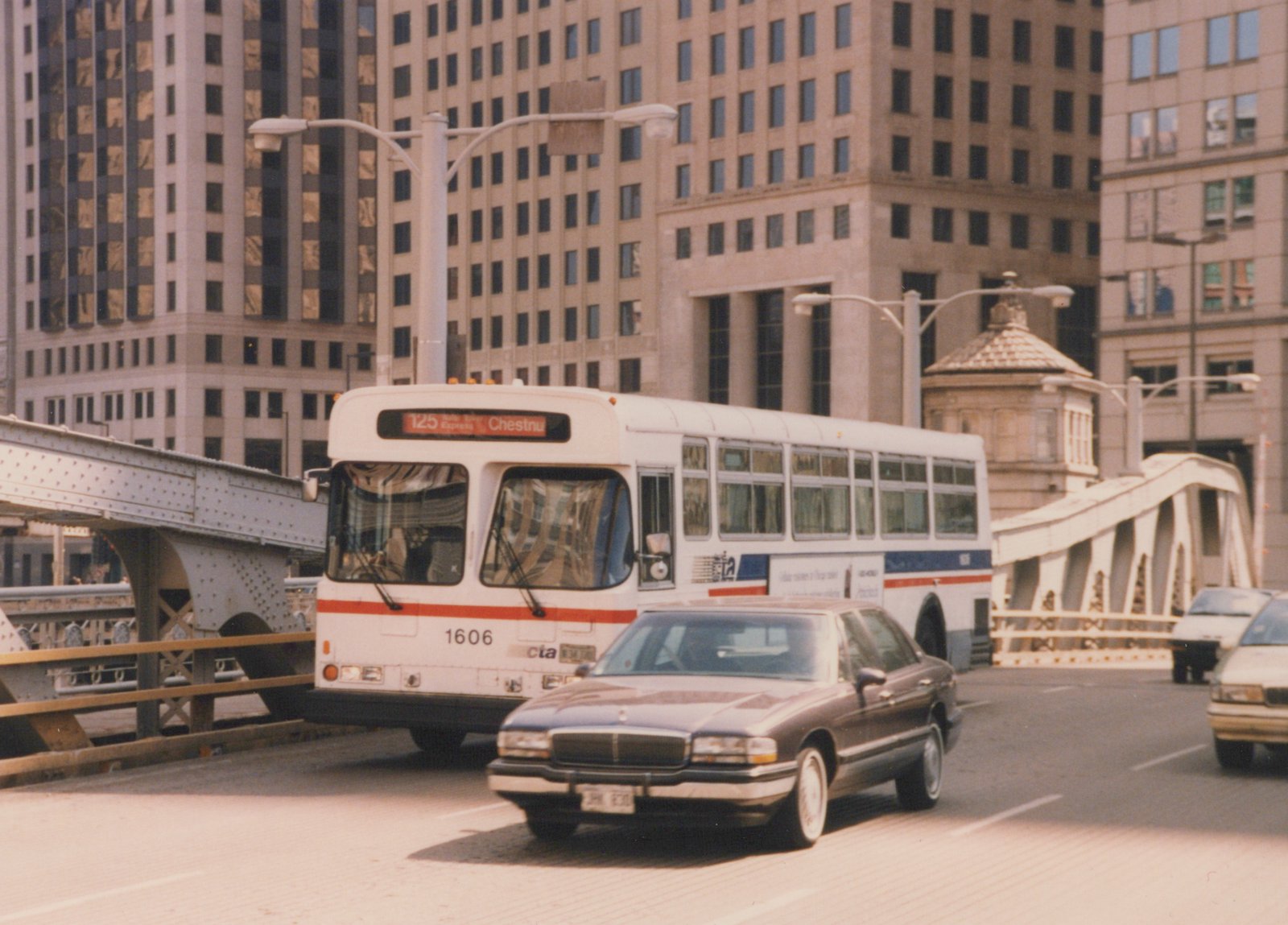 A white Chicago Transit Authority bus with the number 1606 on its front bumper is on a city street, with a brown sedan in front and tall buildings in the background.