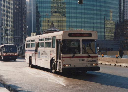 A white CTA bus with the number 1600 on the front drives over a bridge in a city with tall buildings in the background.