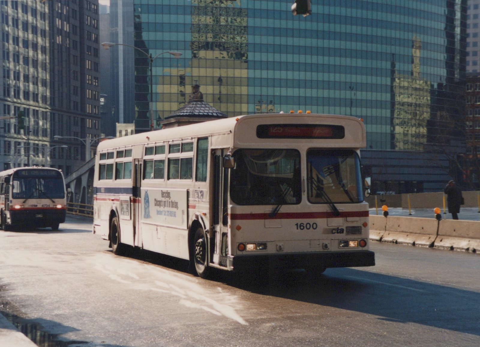 A white CTA bus with the number 1600 on the front drives over a bridge in a city with tall buildings in the background.