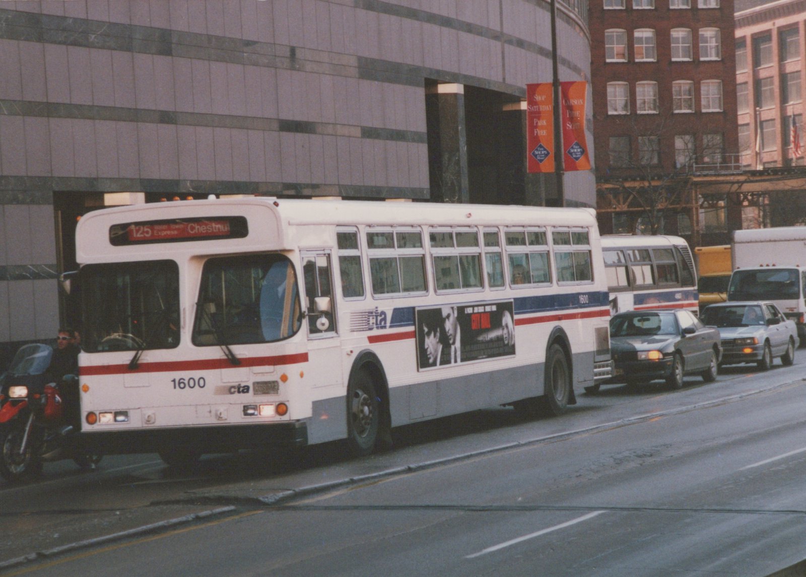 A white Chicago Transit Authority bus, number 1600, is shown on a city street with a '125 Water Tower Express Chestnut' sign on its front, surrounded by cars and a motorcycle.