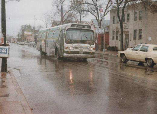 A white and green Chicago Transit Authority bus, number 1514, is driving down a wet street with a police car and a newspaper box nearby.