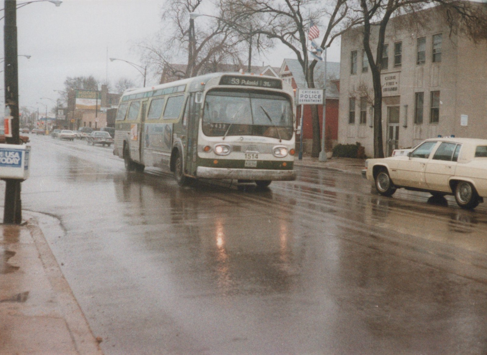 A white and green Chicago Transit Authority bus, number 1514, is driving down a wet street with a police car and a newspaper box nearby.