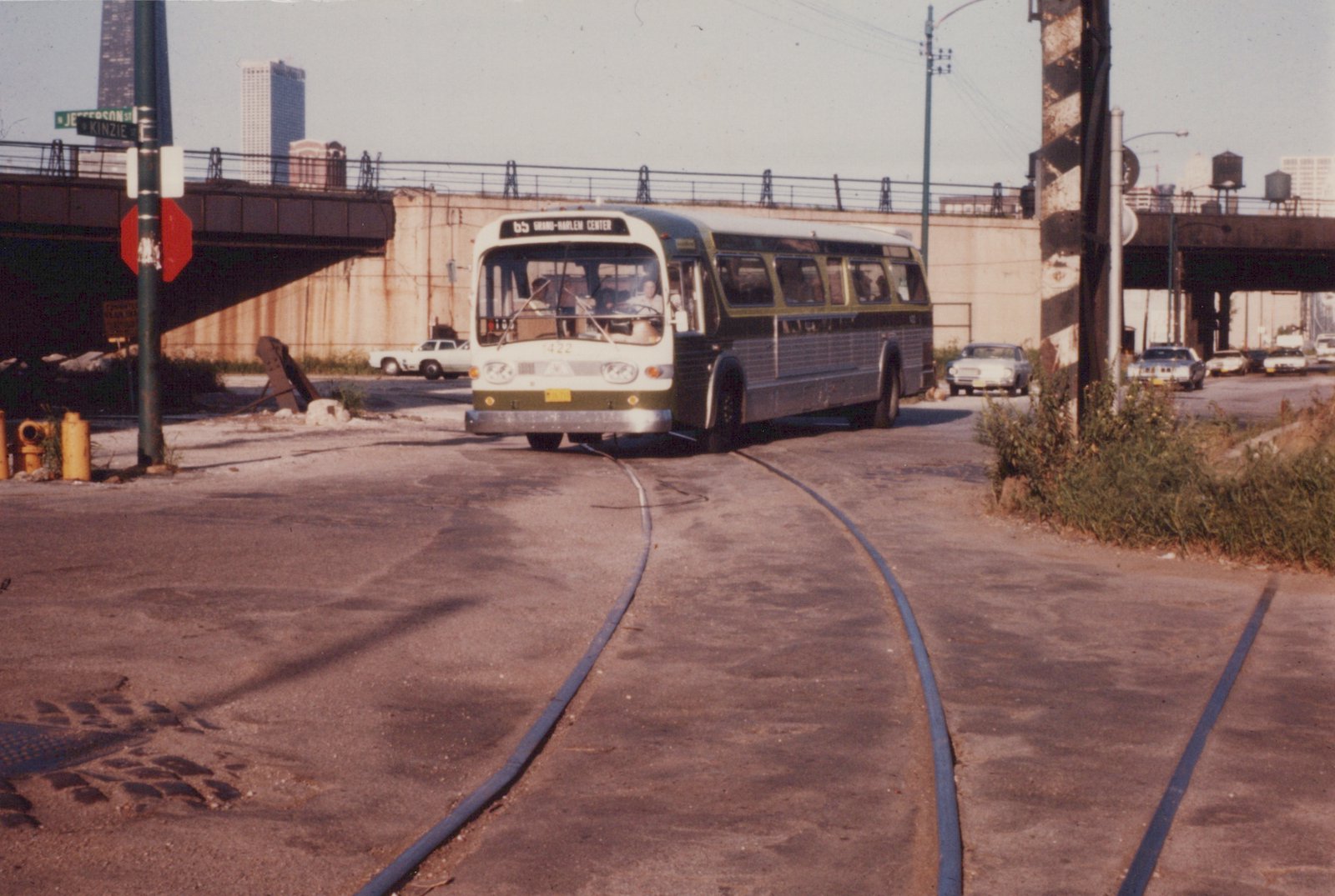 A green and cream CTA bus with the number 1422 is driving on a street next to train tracks, with a street sign for Jefferson and Kinzie Streets visible and a bridge in the background.