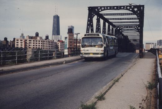 A white and green CTA bus, number 1401, driving over a black metal bridge in Chicago. The Hancock Building is visible in the skyline.