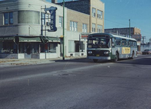 A green and cream transit bus with the number 3094 on its front is turning at a city street corner next to a building with a sign for M. Becker Furs.