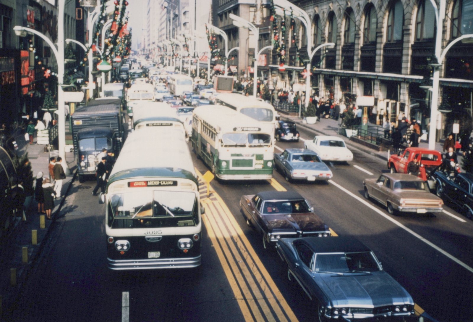 A high-angle view of a Chicago street in winter, featuring a green and white CTA bus with the number 3095 on its front, surrounded by other vintage cars and buses, with festive Christmas decorations on the lampposts.