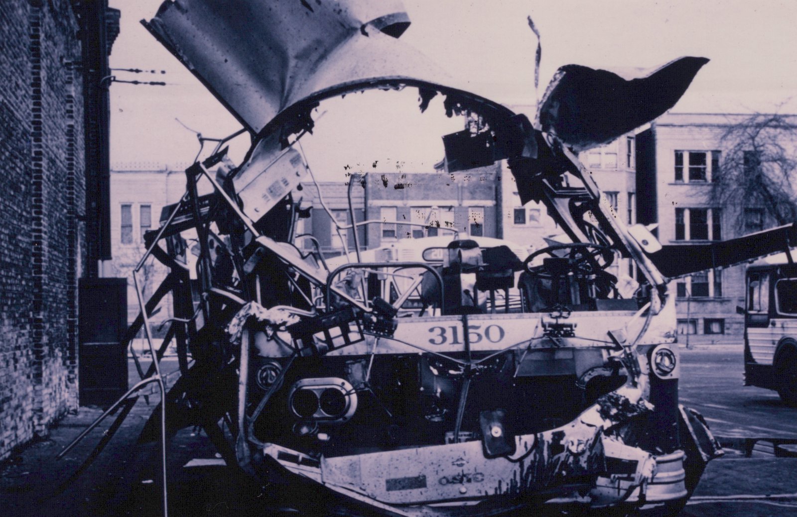 A black and white photo shows the heavily damaged front of a bus with the number 3150, its roof torn off and interior exposed, parked next to a brick building.