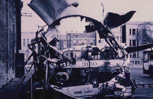 A black and white photo shows the heavily damaged front of a bus with the number 3150, its roof torn off and interior exposed, parked next to a brick building.