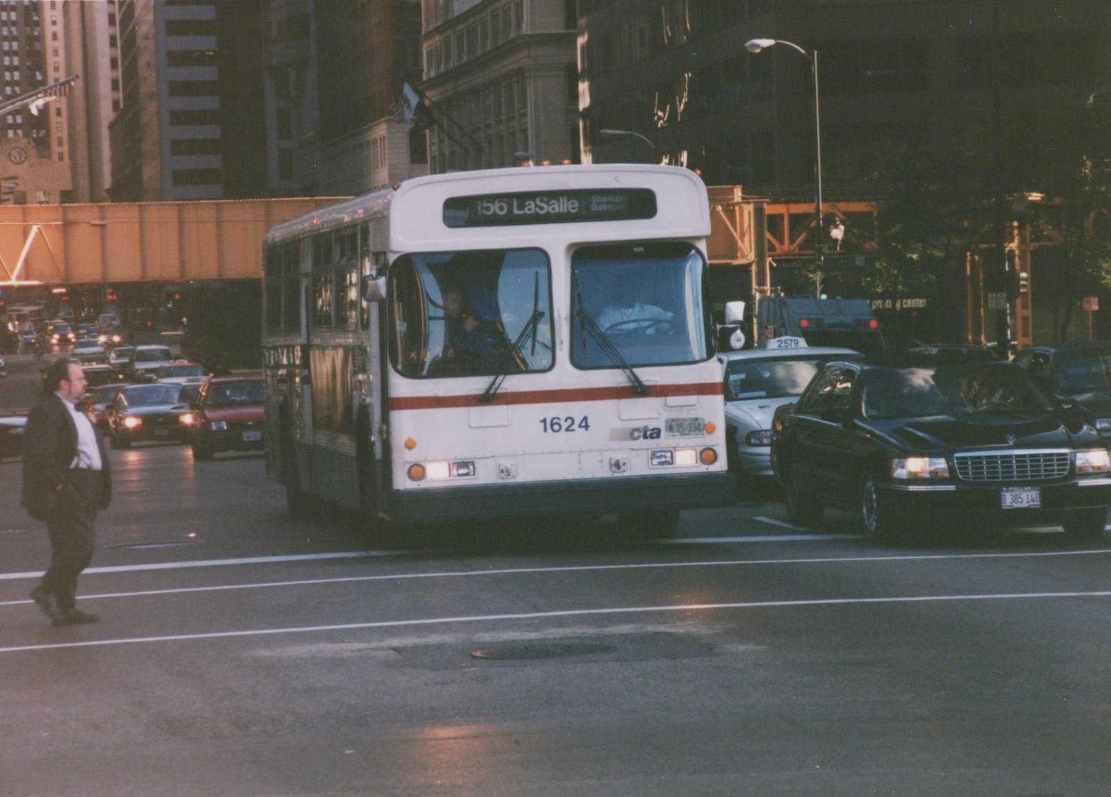 A white and red Chicago Transit Authority bus, fleet number 1624, is stopped in city traffic with its headlights on, surrounded by cars and a pedestrian crossing the street.