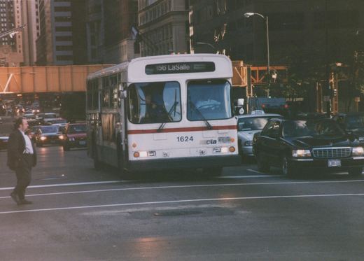 A white and red Chicago Transit Authority bus, fleet number 1624, is stopped in city traffic with its headlights on, surrounded by cars and a pedestrian crossing the street.