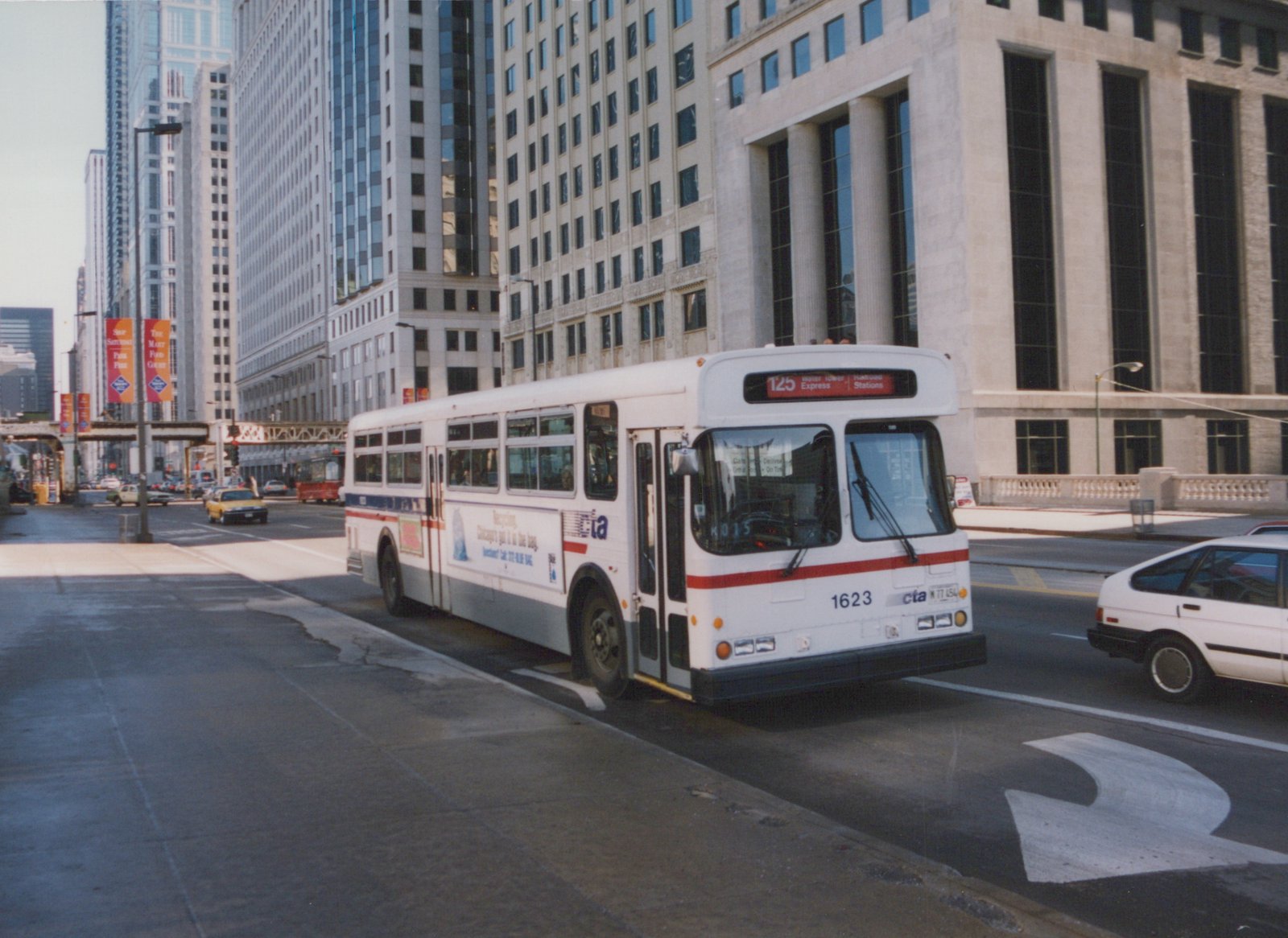 A white Chicago Transit Authority bus with the number 1623 is driving down a city street lined with tall buildings, with other cars visible nearby.