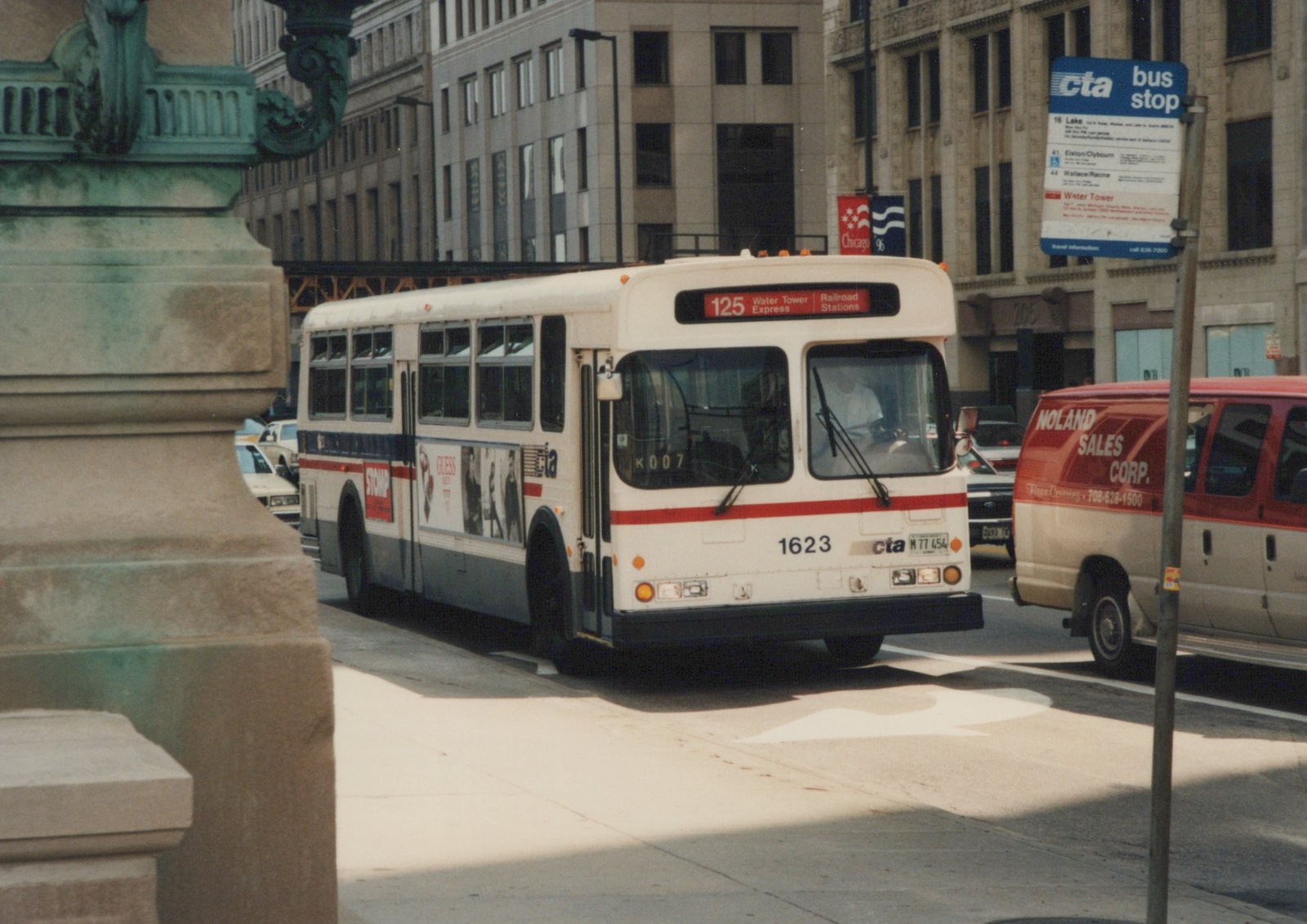A white CTA bus with red and blue stripes, number 1623, is stopped at a bus stop on a sunny city street, with a red and white van and tall buildings in the background.