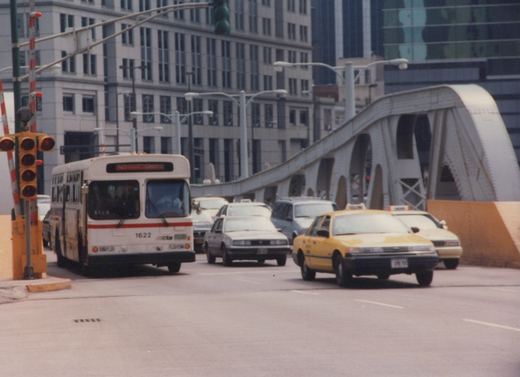 A white and red CTA bus with the number 1622 is stopped at an intersection with several cars and a yellow taxi, with a large metal bridge in the background.