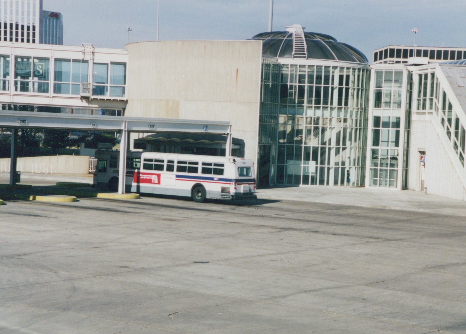 A white bus with red and blue stripes, number 1621, is parked at a bus terminal with a distinctive glass-domed building in the background.