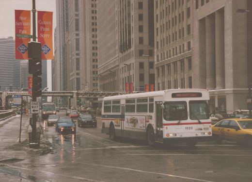 A white and blue Chicago Transit Authority bus, number 1620, drives on a wet city street lined with tall buildings and banners, with other vehicles and a yellow taxi nearby.