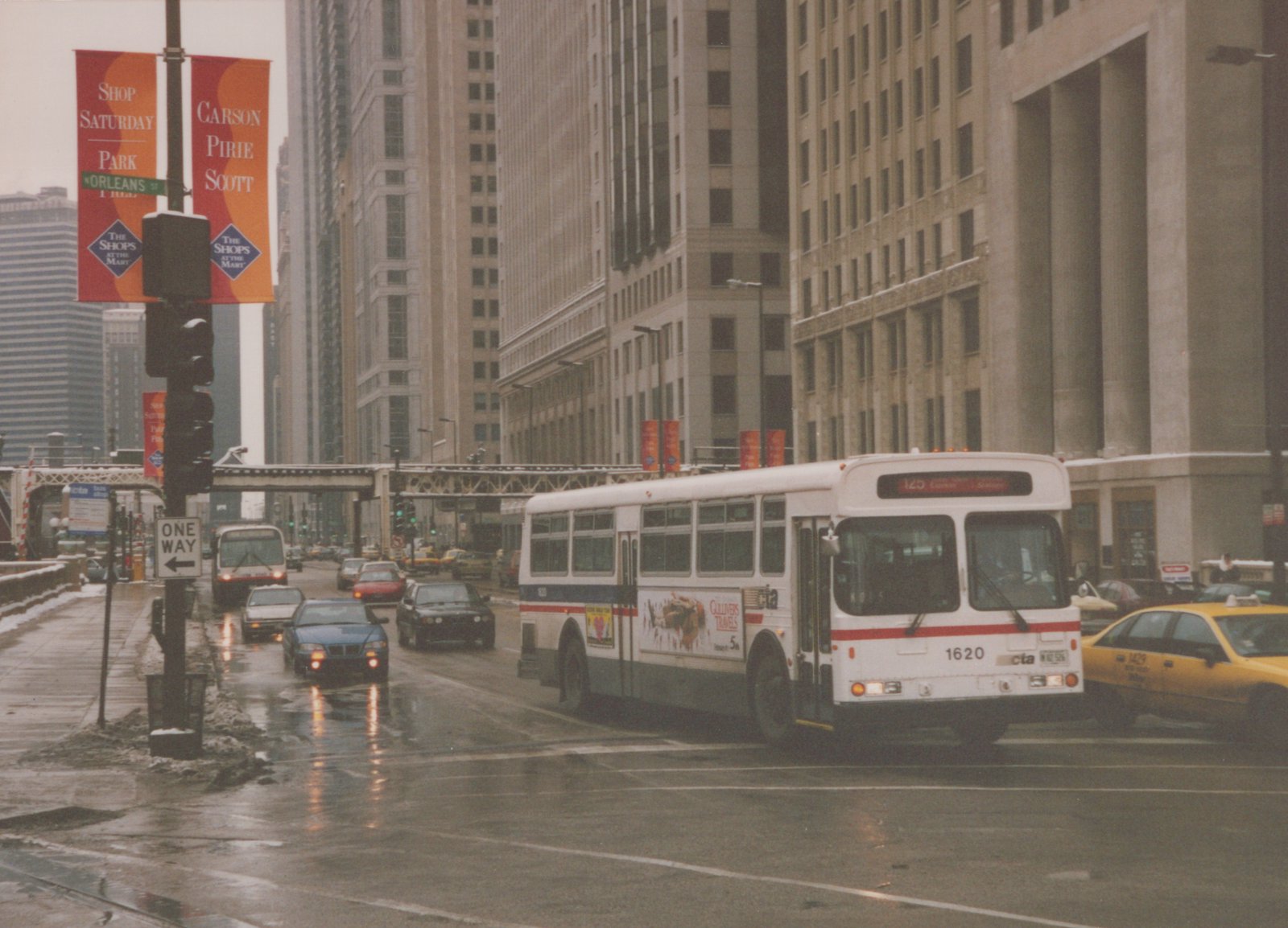 A white and blue Chicago Transit Authority bus, number 1620, drives on a wet city street lined with tall buildings and banners, with other vehicles and a yellow taxi nearby.