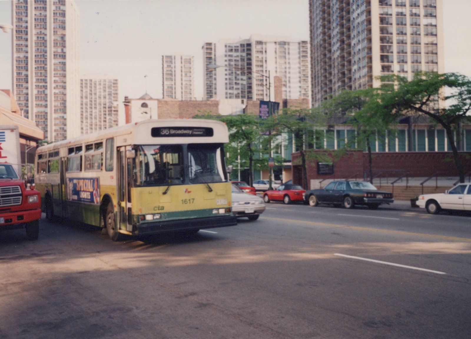 A green and cream CTA bus with the number 1617 and route 36 Broadway is on a city street with tall buildings and several cars from the 1980s in the background.