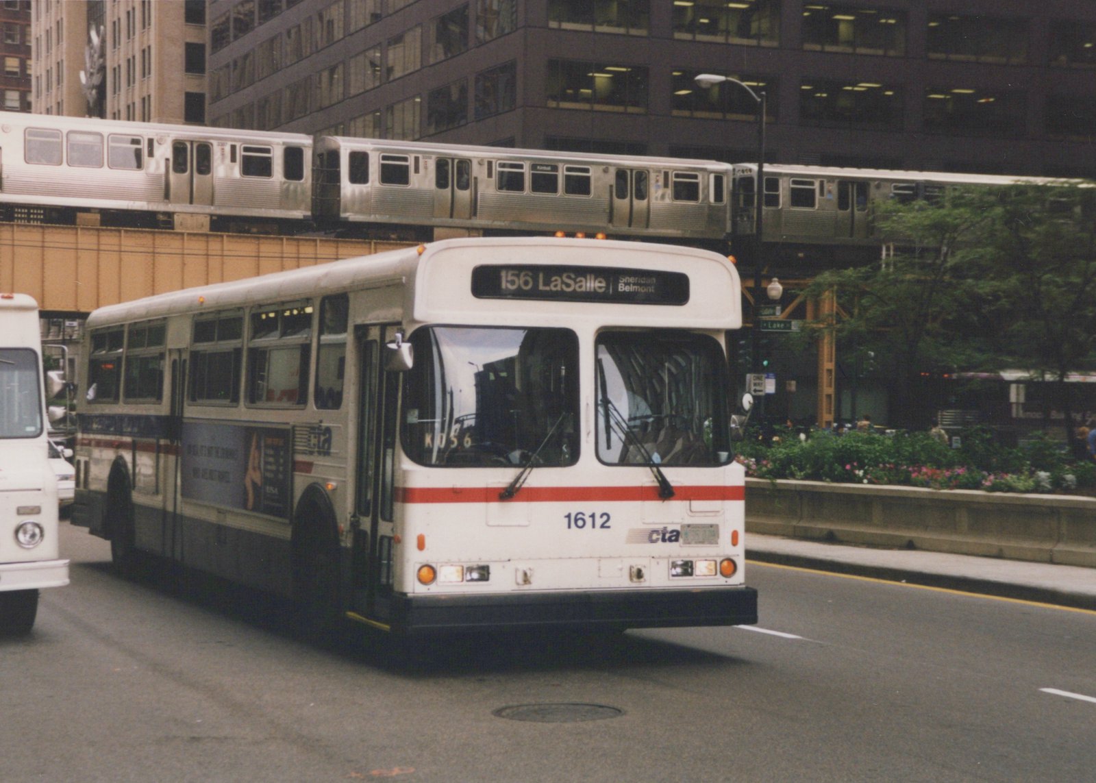 A white CTA bus with red and blue stripes is on a city street, with a silver 'L' train on an elevated track above it.