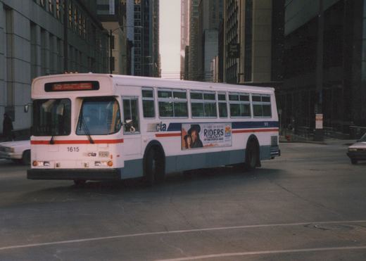 A white, blue, and red CTA bus with fleet number 1615 is seen from a three-quarter front view, driving down a city street lined with tall buildings.