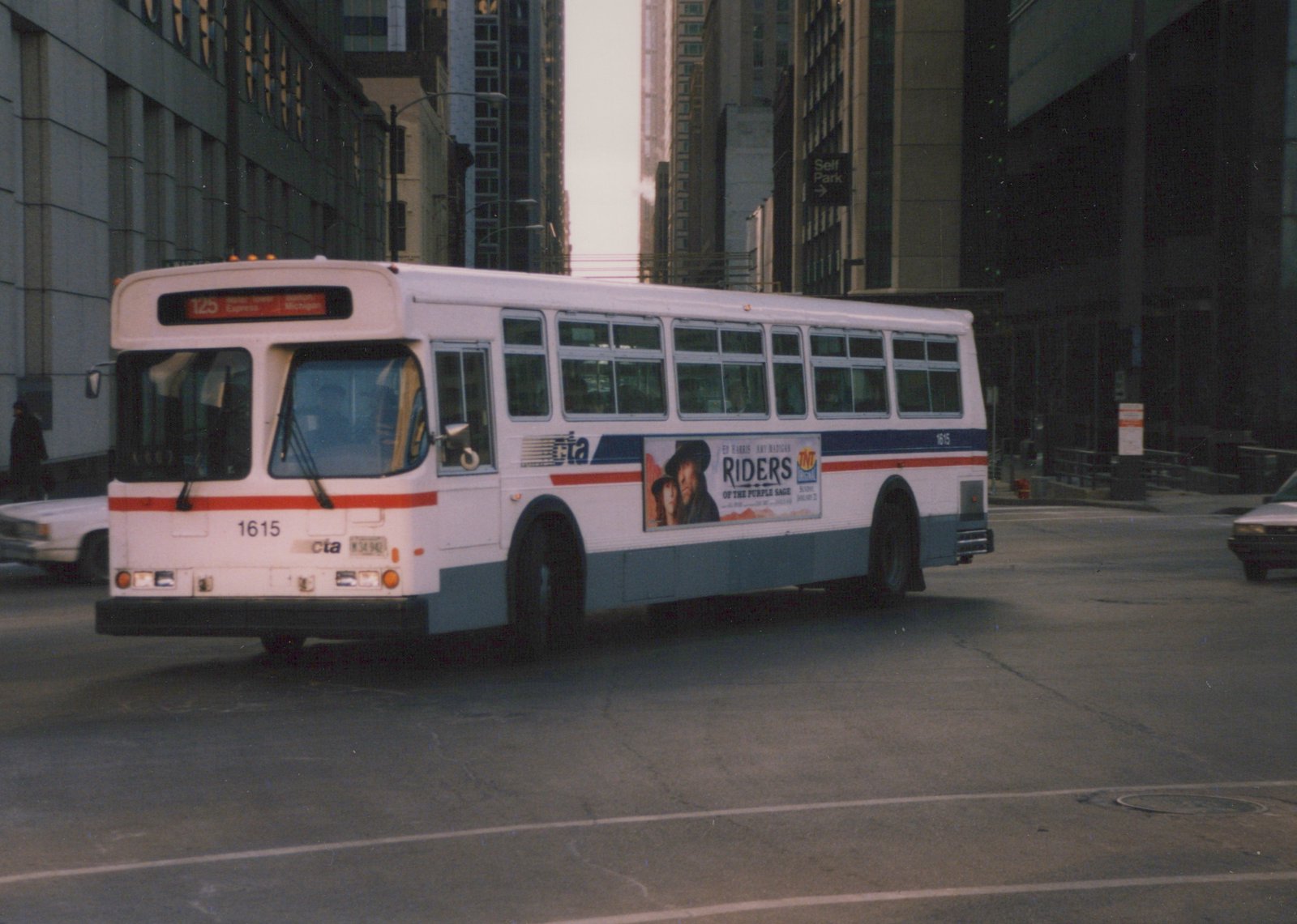 A white, blue, and red CTA bus with fleet number 1615 is seen from a three-quarter front view, driving down a city street lined with tall buildings.