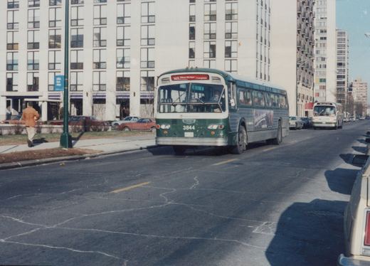 A green and white CTA bus with the number 3844 is driving down a city street with tall buildings in the background, and another bus is visible behind it.