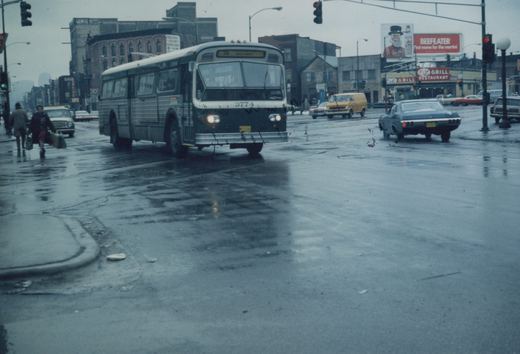 A green and white Chicago Transit Authority bus, number 3774, drives on a wet street with pedestrians and other cars, under overcast skies with rain.
