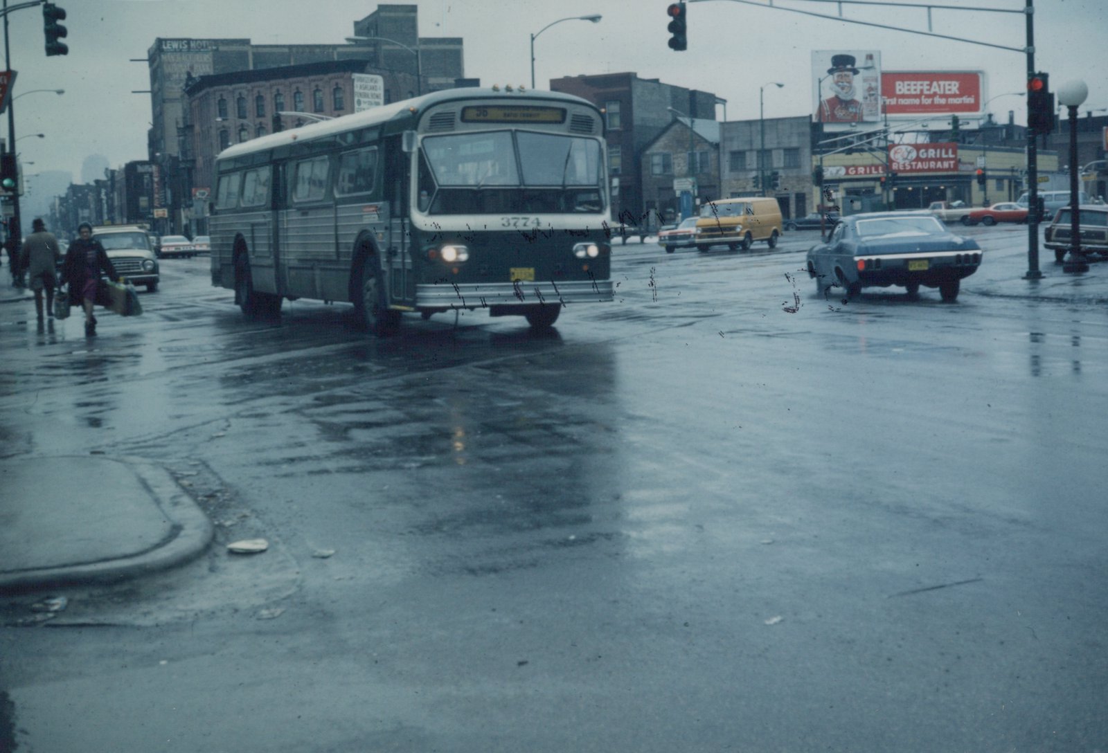A green and white Chicago Transit Authority bus, number 3774, drives on a wet street with pedestrians and other cars, under overcast skies with rain.