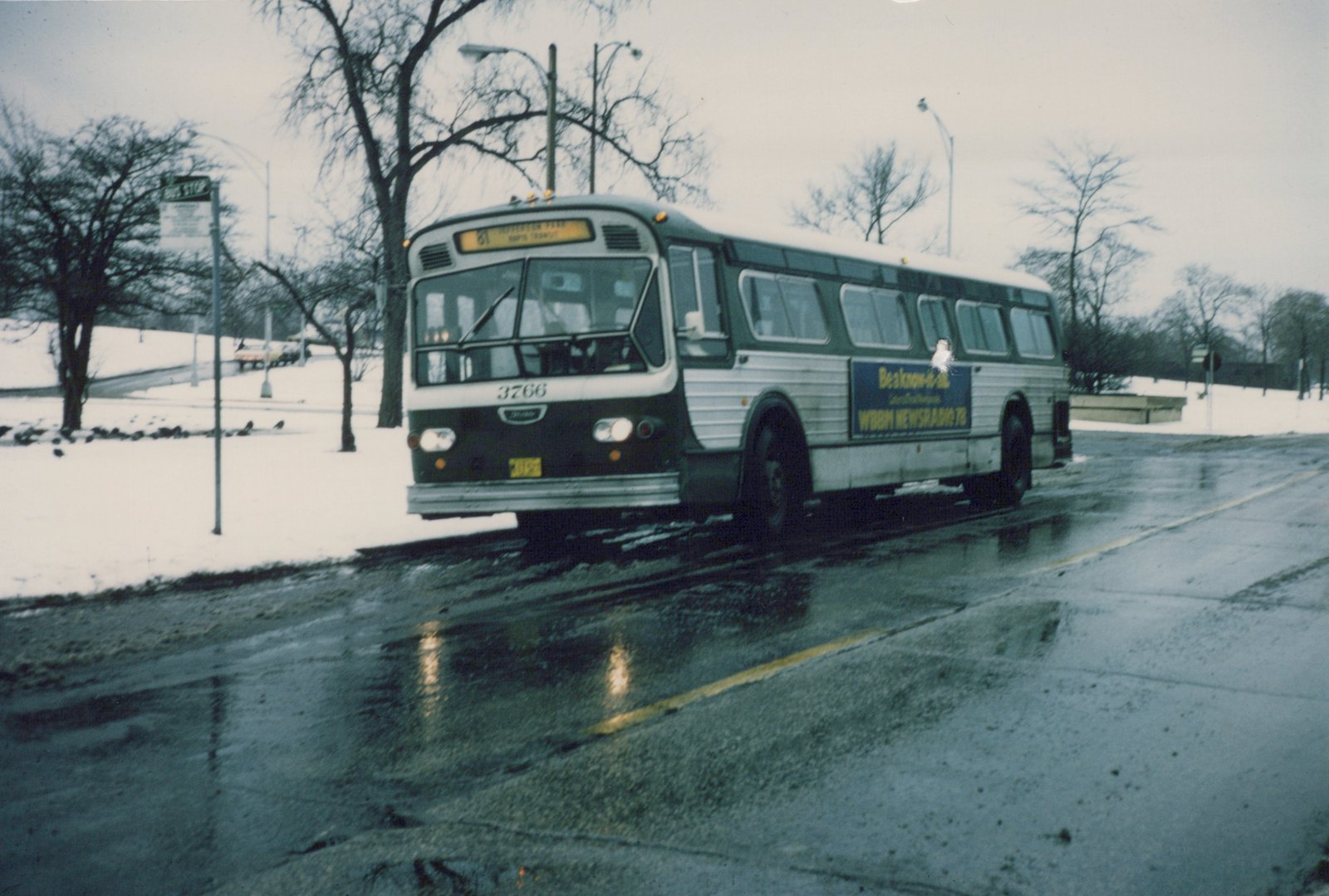 A white and green CTA bus is stopped at a snow-covered bus stop on a wet street, with bare trees and a cloudy sky in the background.