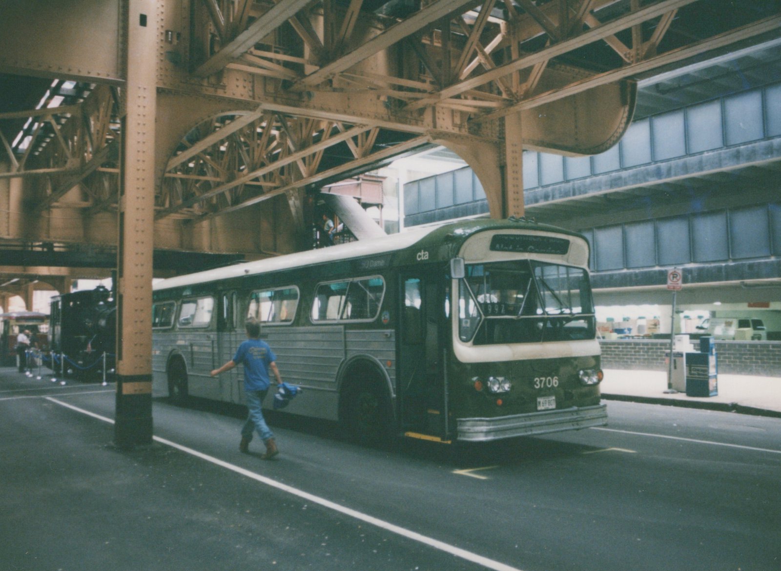 A green and cream CTA bus numbered 3706 is parked under a large steel elevated train structure. A person walks in front of the bus, and a black steam locomotive is visible in the background.