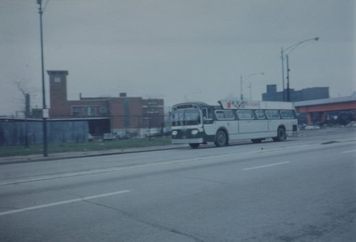 A green and white motor bus with the number 4[?]71 is driving down a wide, overcast city street next to a brick building and under an elevated structure.