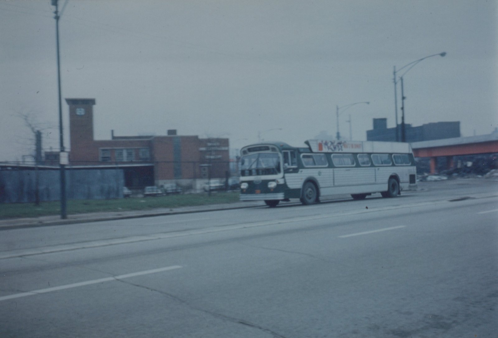 A green and white motor bus with the number 4[?]71 is driving down a wide, overcast city street next to a brick building and under an elevated structure.