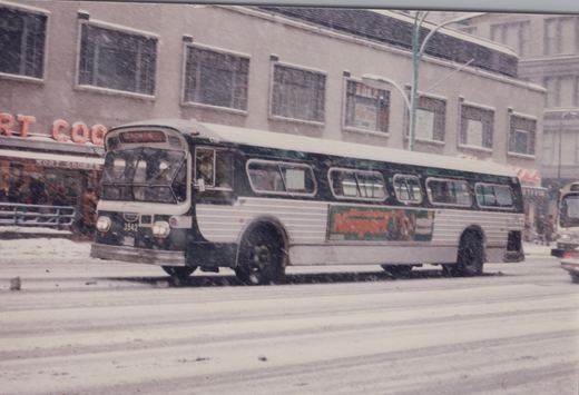 A green and white Chicago Transit Authority bus drives on a snow-covered street, with storefronts in the background and another bus partially visible behind it.