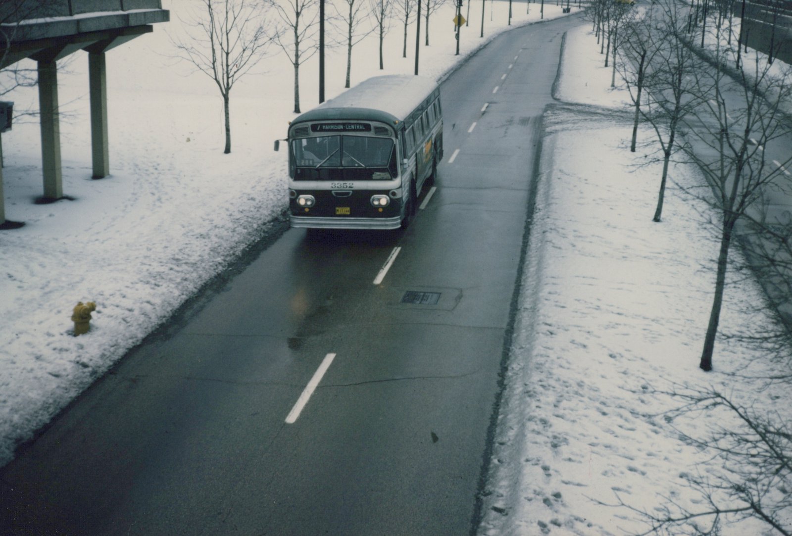 A green and white bus with the number 3352 and destination 'HARRISON-CENTRAL' drives on a wet, snow-lined road under an overpass.