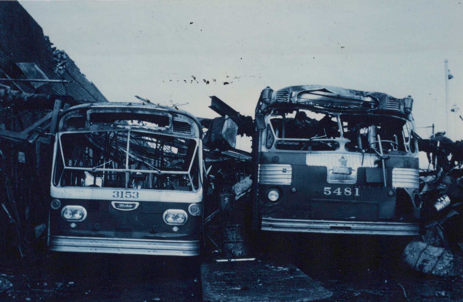 Two heavily damaged and burned-out buses with fleet numbers 3153 and 5481, showing signs of being in a scrapyard.