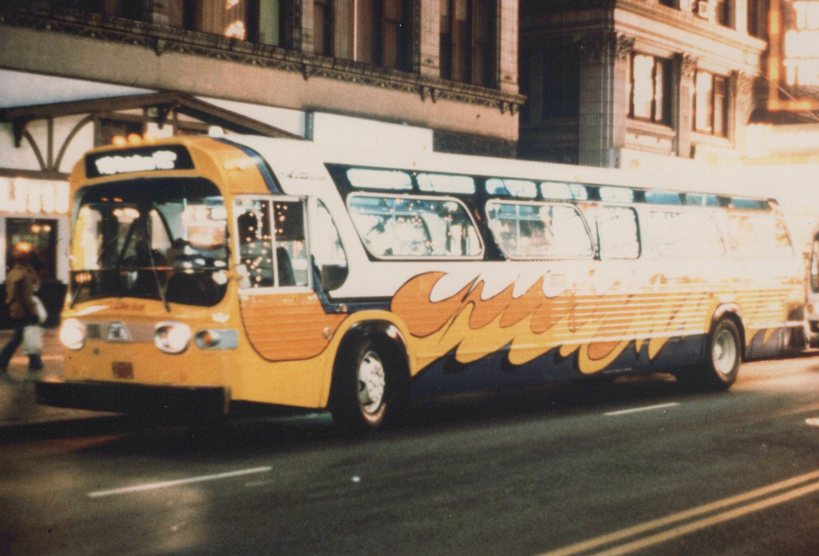 A yellow, white, and blue bus with flame graphics is driving down a city street with buildings in the background.