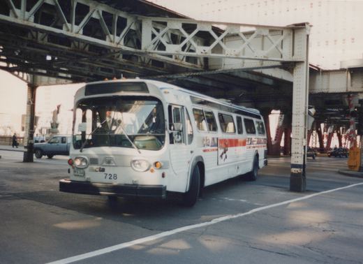 A white CTA bus, number 728, is shown from a three-quarter front view as it drives on a city street beneath an elevated steel truss structure on a clear day.