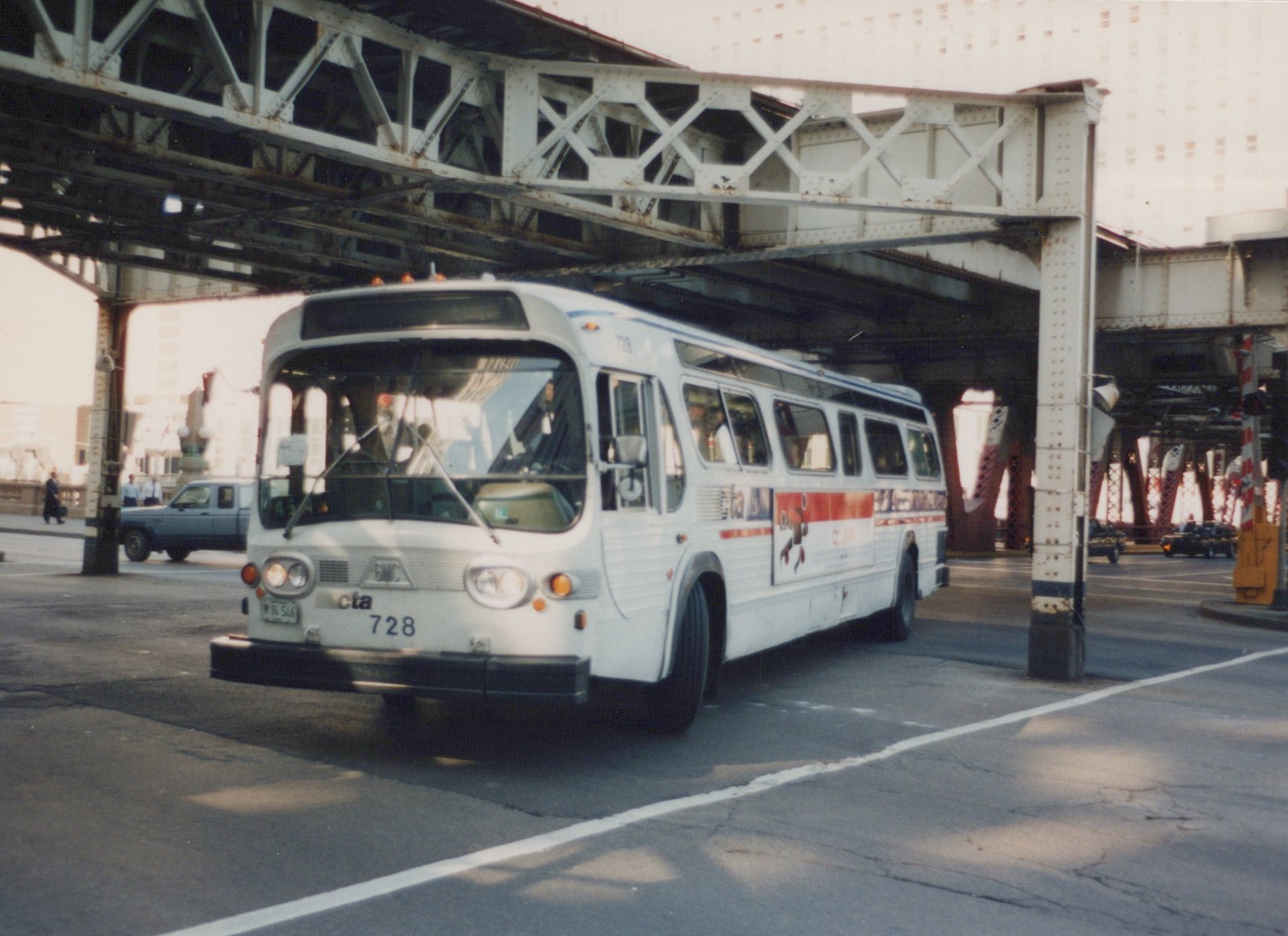 A white CTA bus, number 728, is shown from a three-quarter front view as it drives on a city street beneath an elevated steel truss structure on a clear day.