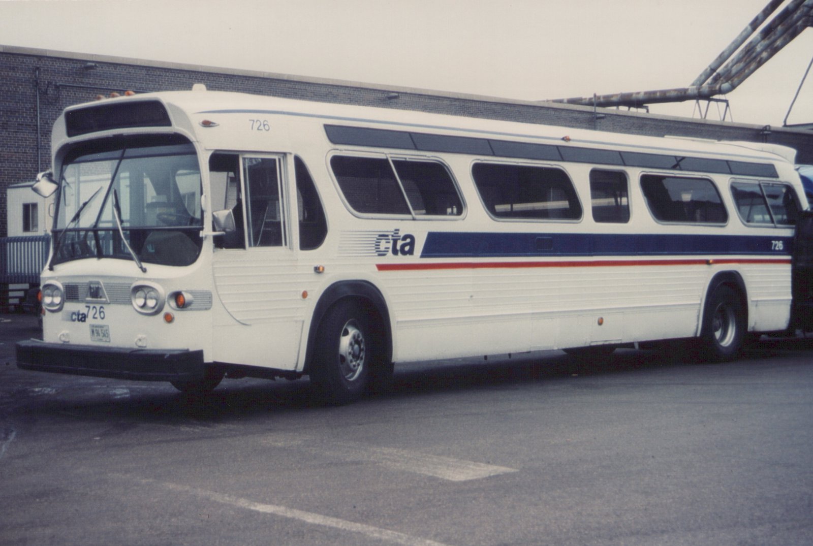 A white CTA bus with blue and red stripes, number 726, is parked in a lot with a brick building behind it.
