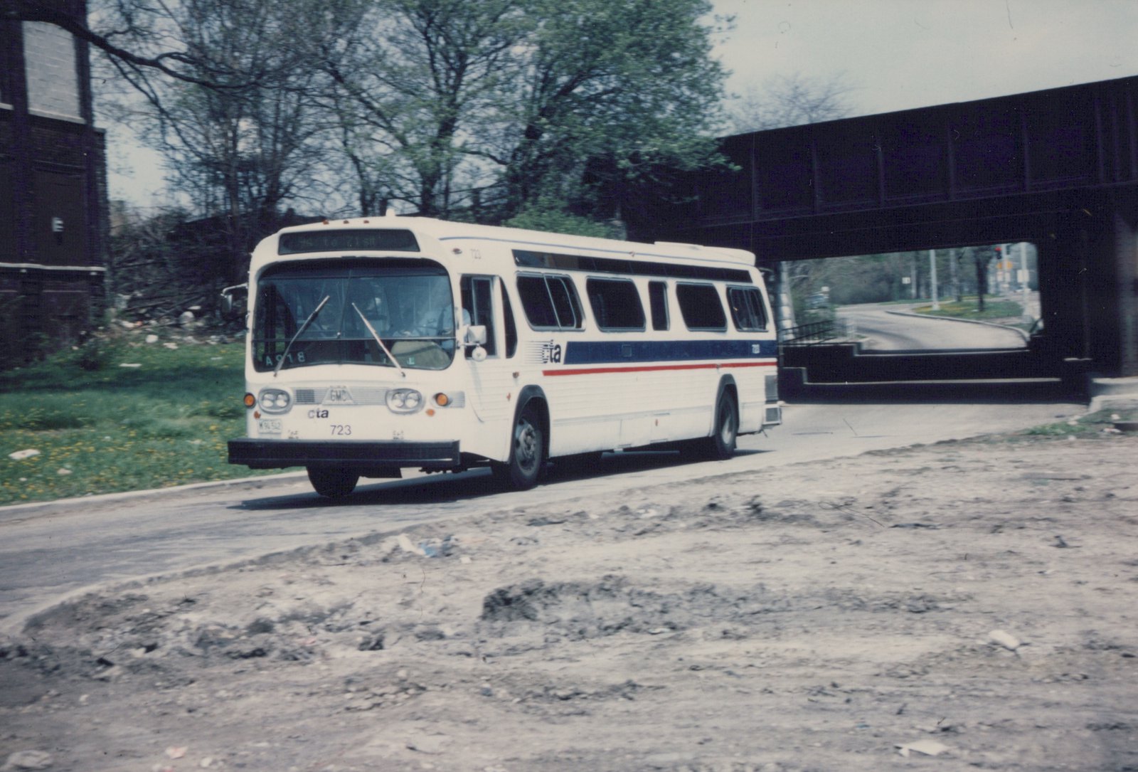 A white CTA bus with blue and red stripes, number 723, is driving on a paved road with a bridge in the background and a grassy area to the side.