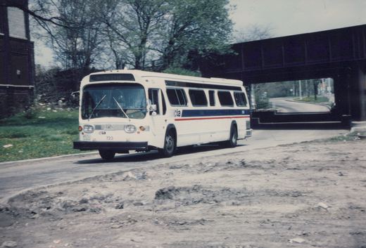 A white CTA bus with blue and red stripes, number 723, is driving on a paved road with a bridge in the background and a grassy area to the side.