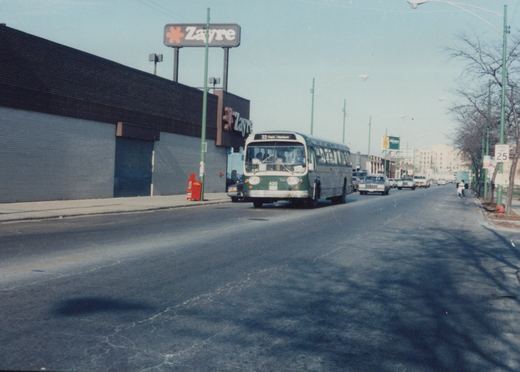 A green and white city bus drives down a street past a Zayre store, with cars and bare trees lining the road.