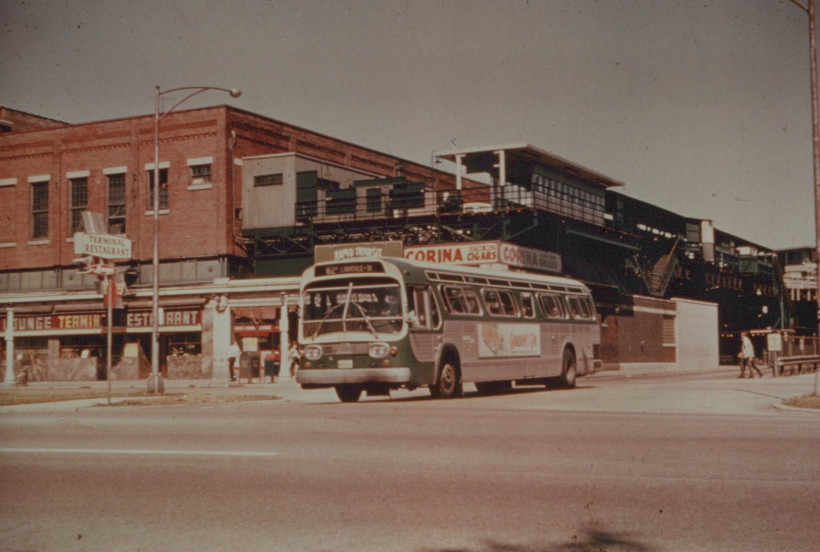 A green and cream Chicago Transit Authority bus with the number 3077 travels on a street under an elevated train structure, with a 'Terminal Restaurant' and 'Corina Cigars' signs visible nearby.