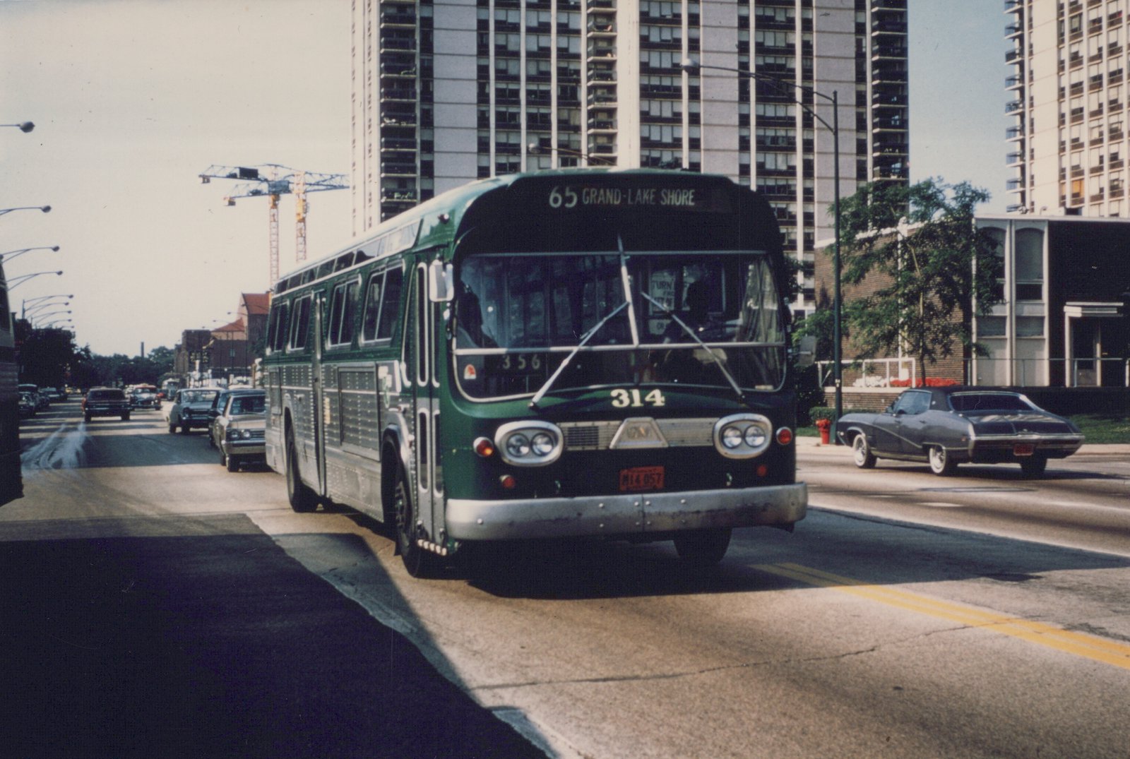A green CTA bus with the number 314 and destination '65 Grand-Lake Shore' is driving down a city street with several cars and tall buildings in the background on a sunny day.