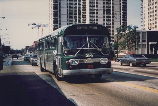 A green CTA bus with the number 314 and destination '65 Grand-Lake Shore' is driving down a city street with several cars and tall buildings in the background on a sunny day.