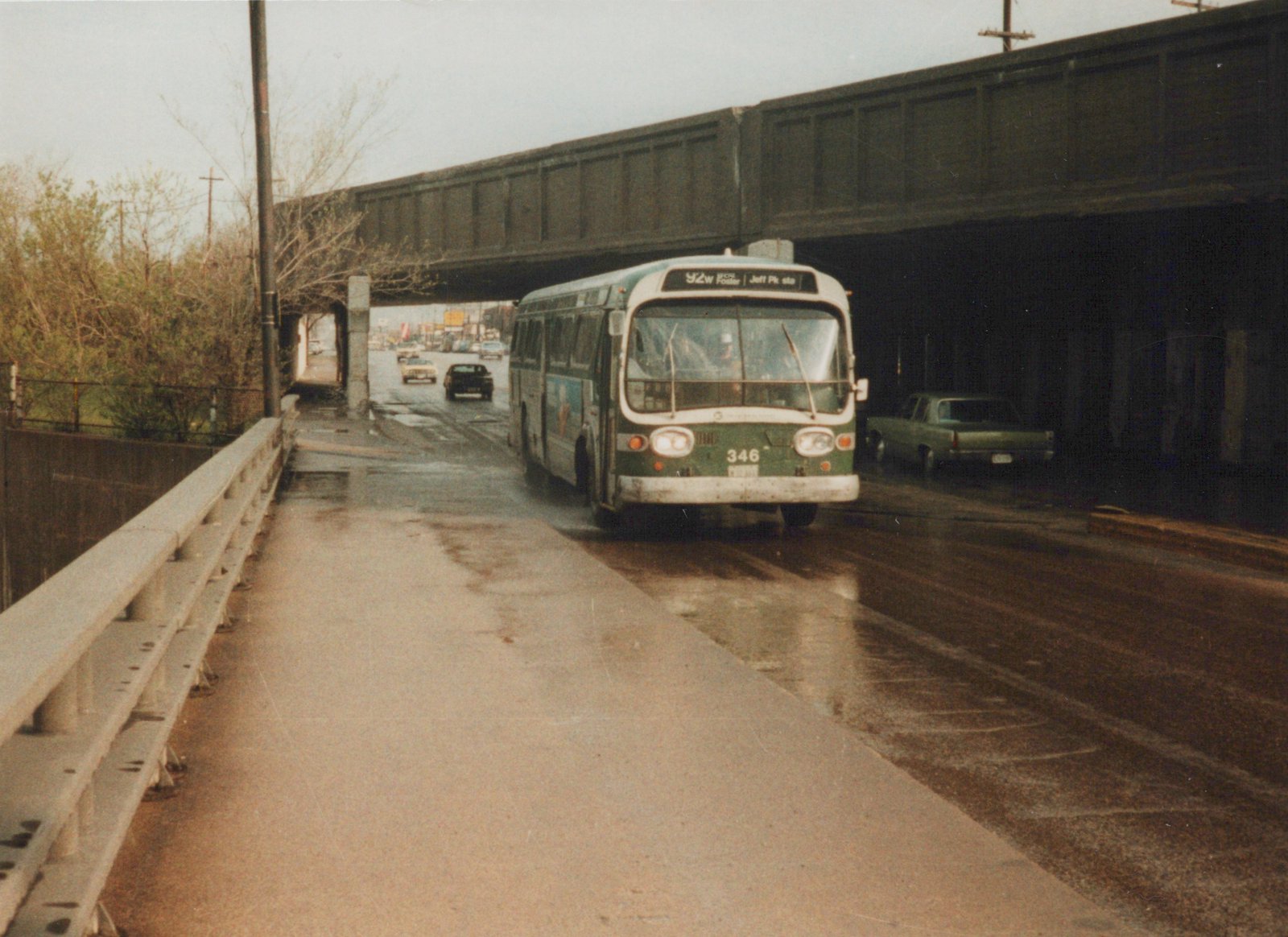 A green and cream CTA bus, number 346, drives on a rain-slicked street under a large bridge, with cars visible in the background.