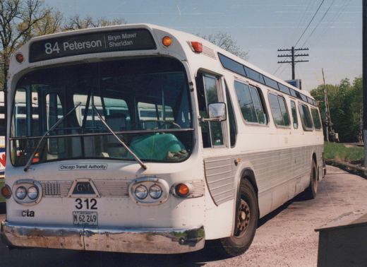 A white and silver CTA bus number 312 is parked on a city street beside railroad tracks under a clear blue sky, with its destination sign reading "84 Peterson".