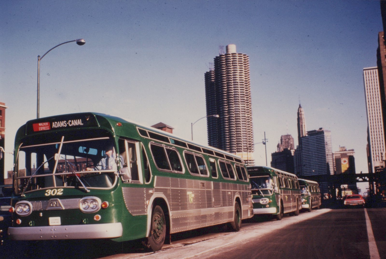 A green and silver Chicago Transit Authority bus, number 302, is shown from a three-quarter front view on a city street with the Marina City towers in the background.