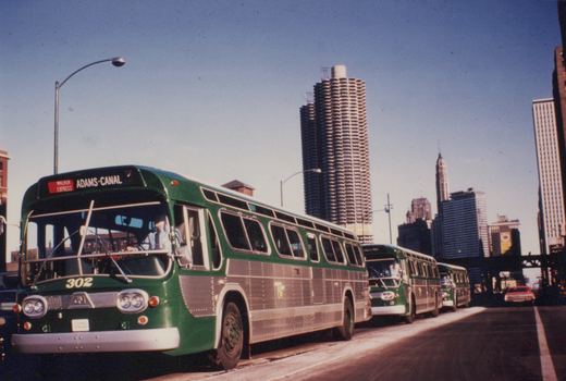 A green and silver Chicago Transit Authority bus, number 302, is shown from a three-quarter front view on a city street with the Marina City towers in the background.